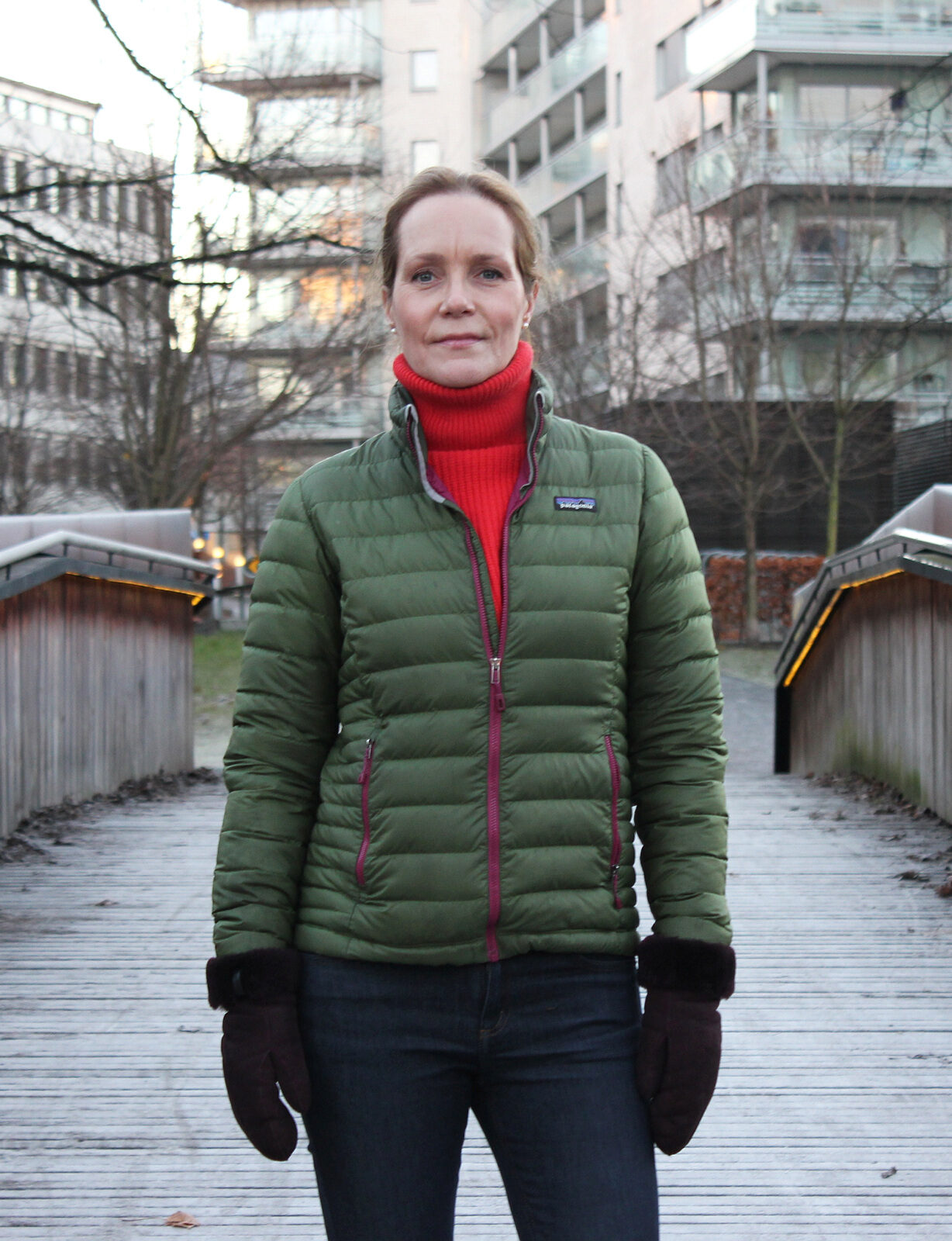 bilde av Hanna Geiran kledt i grønn boblejakke, rød genser og votter. Hun står på en frostbelagt trebro ved Akerselva i Oslo.